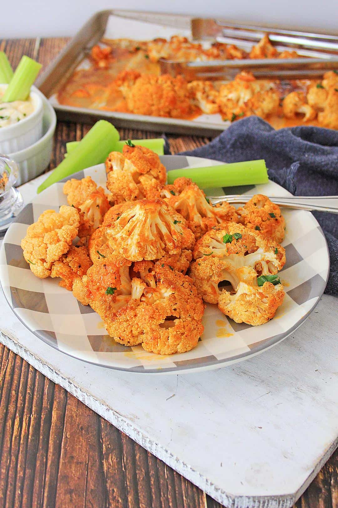 Roasted buffalo cauliflower on a plate with sheet pan in background with tongs, Clery and ranch dip.