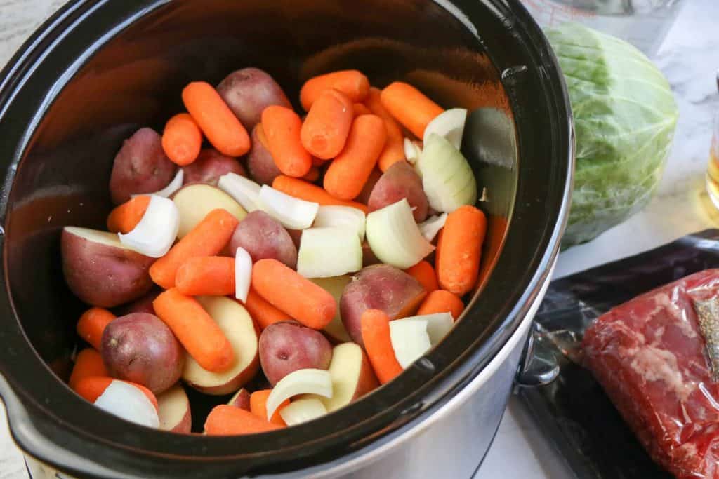 Carrot, baby red potatoes and onions in a slow cooker with brisket beside the slow cooker.