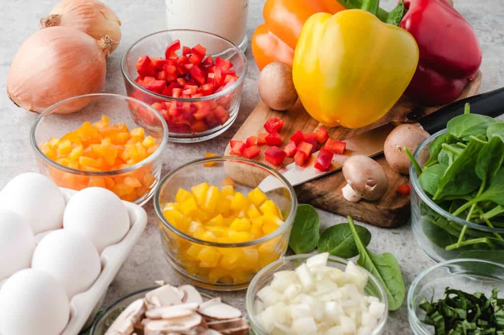 chopped and whole bell peppers, onions, spinach, eggs and mushrooms on an off white counter top
