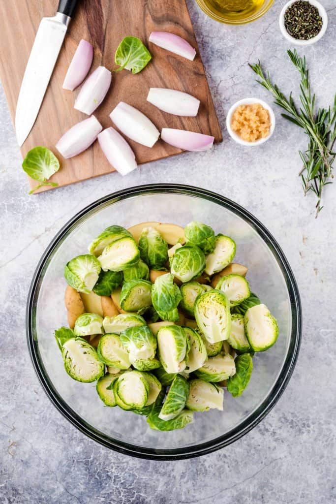 Brussels sprouts and potatoes in a bowl with shallots above them on a cutting board.