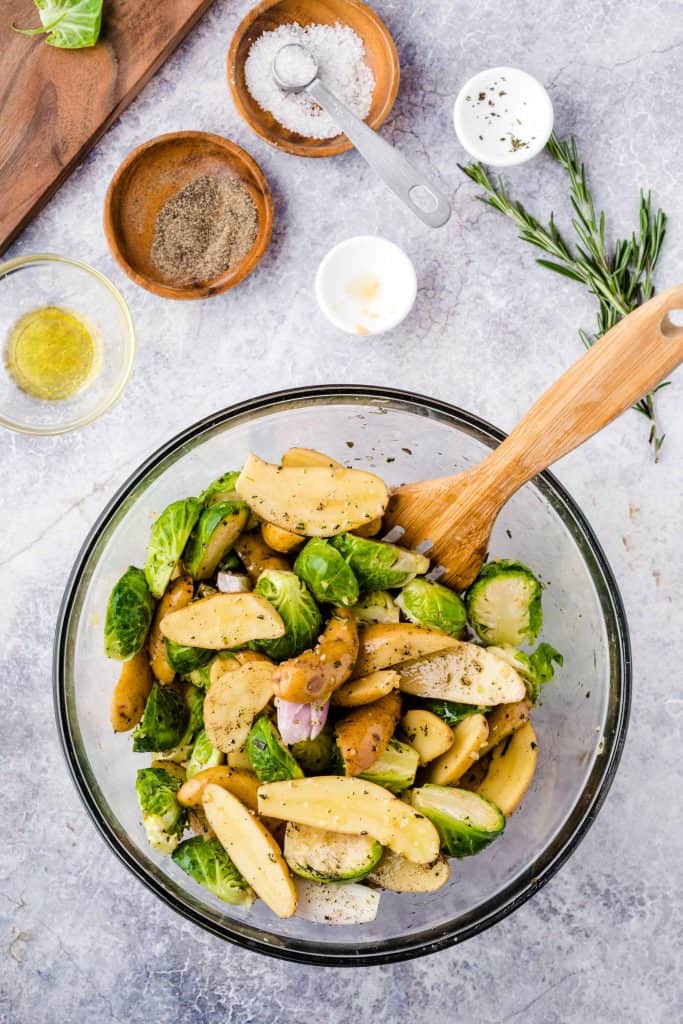 Fingerling potatoes, Brussels sprouts and shallots in a glass bowl with salt, pepper and herbs above it.