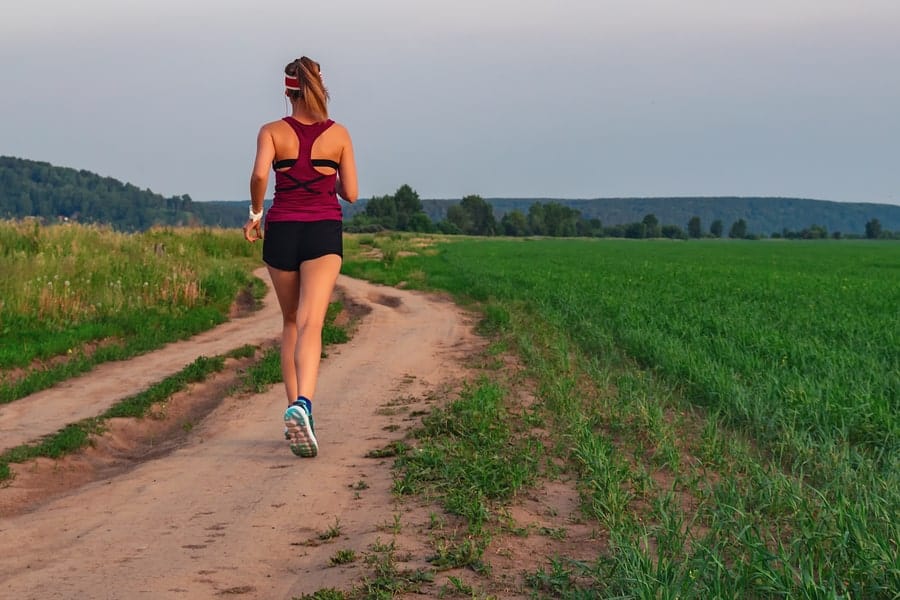 Athletic Blonde Teenage Girl Running At Dirt road In field. Sport girl running outdoor. Young woman running in field. Back view running lass on meadow at sunset. Fitness training.