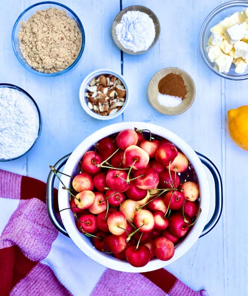 Overhead view of Rainier cherries in a white colander, brown sugar, flour, chopped pecans Arrow Root powder, cinnamon, salt, butter and lemon with a red and white dish towel on the eggs.