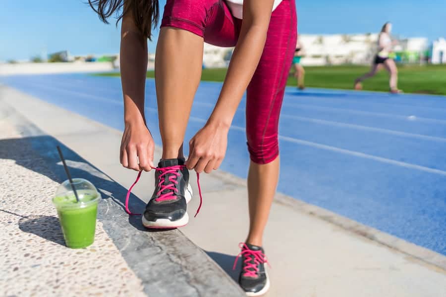 Healthy lifestyle woman runner tying running shoes drinking green smoothie cup juice drink before race workout on run tracks at outdoor stadium. Athlete getting ready for cardio training.