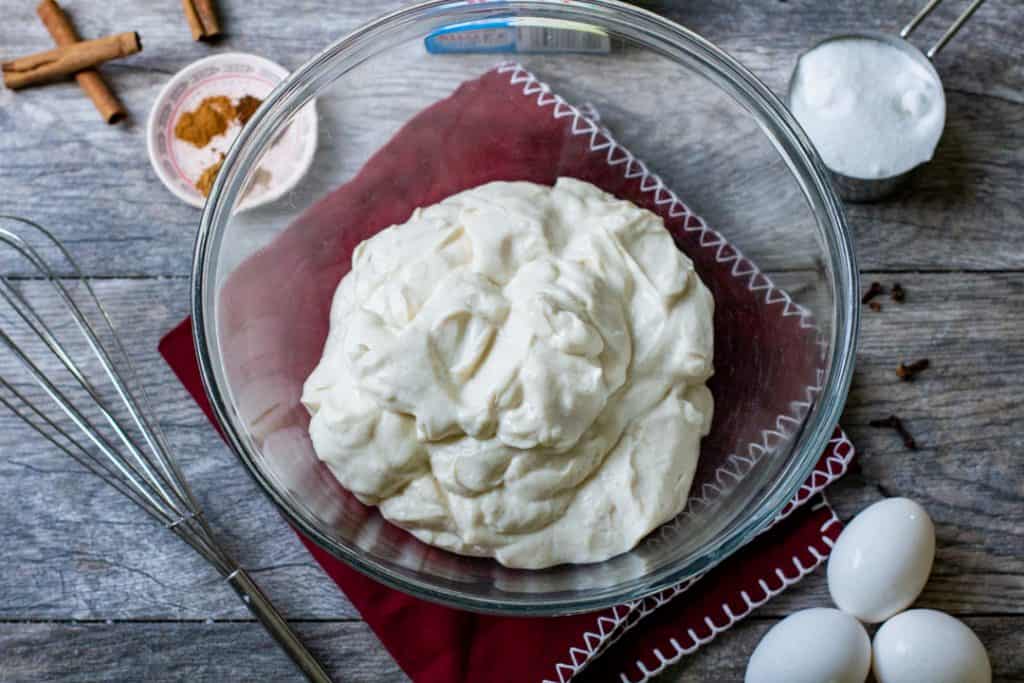 Greek yogurt in a bowl with eggs, cinnamon sticks, and whisk on wood table