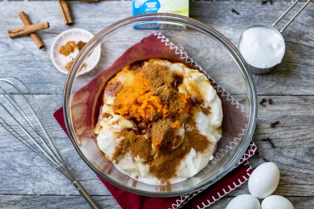 Pumpkin cheesecake bite ingredients in a bowl with cinnamon sticks, eggs and whisk on wood table
