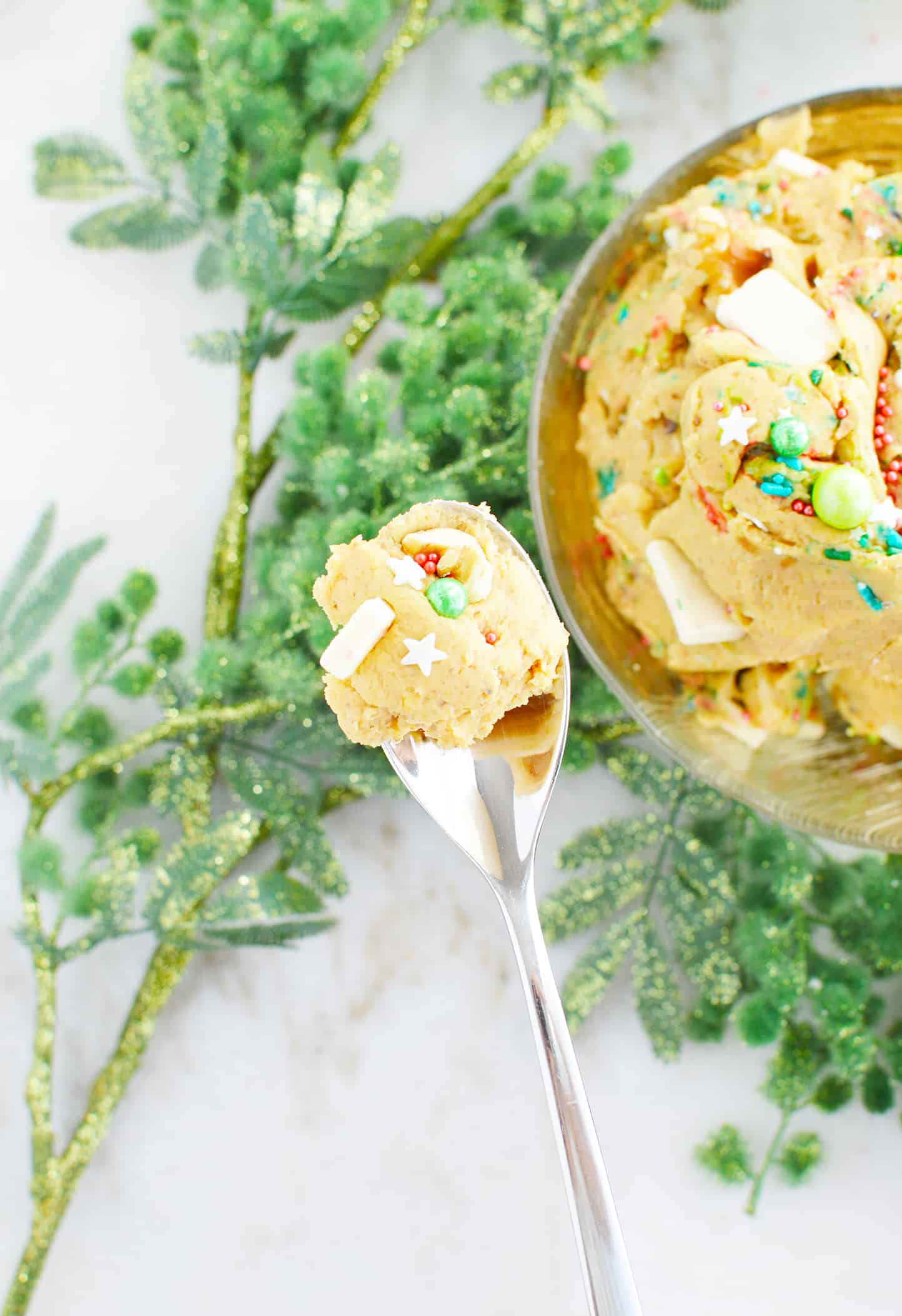 Healthy Edible Cookie Dough on a spoon with evergreen branches and bowl of healthy edible cookie dough below it.