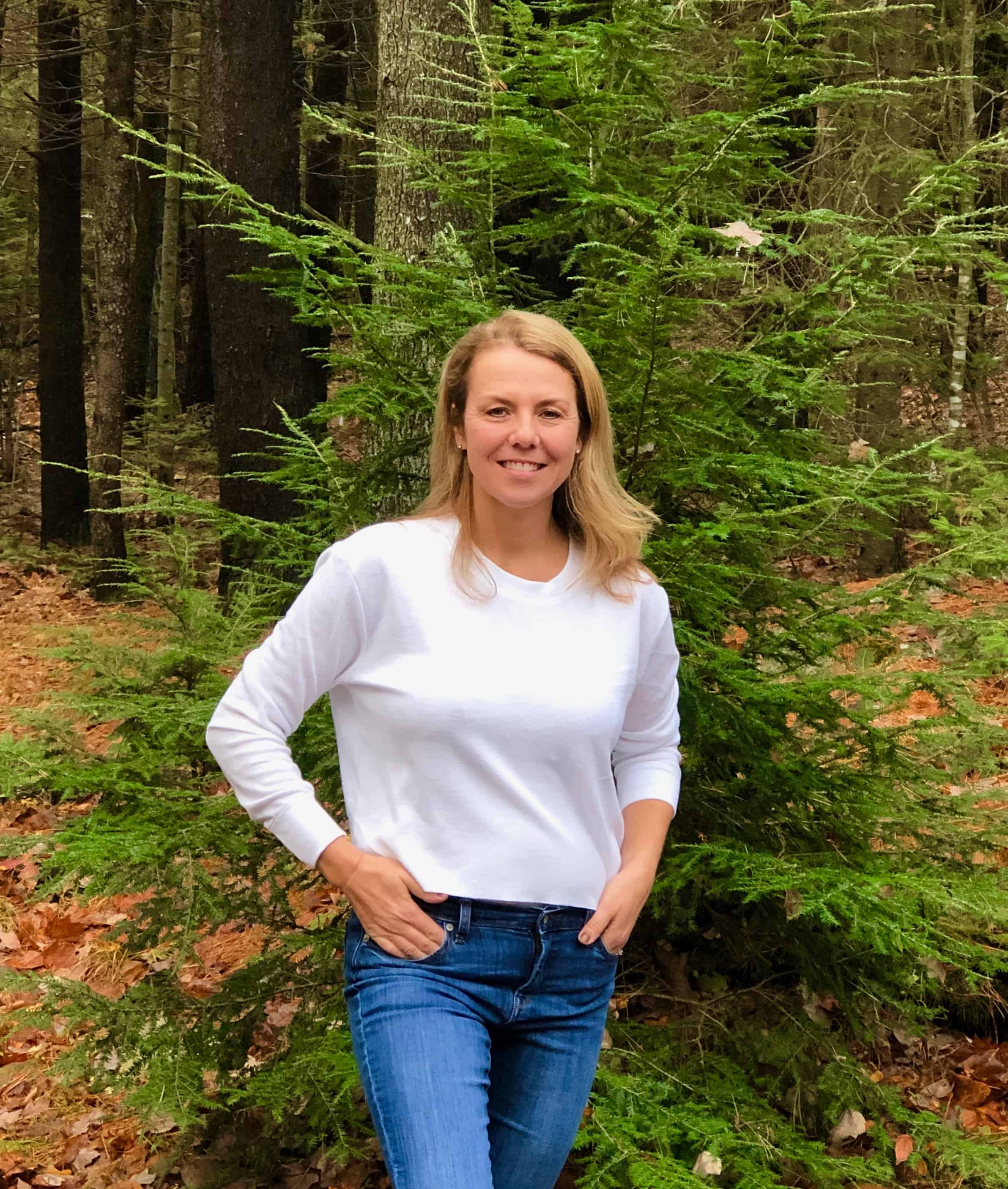 Beautiful blond woman in white thermal crop top and denim jeans standing outside by tree in Maine during the Fall.