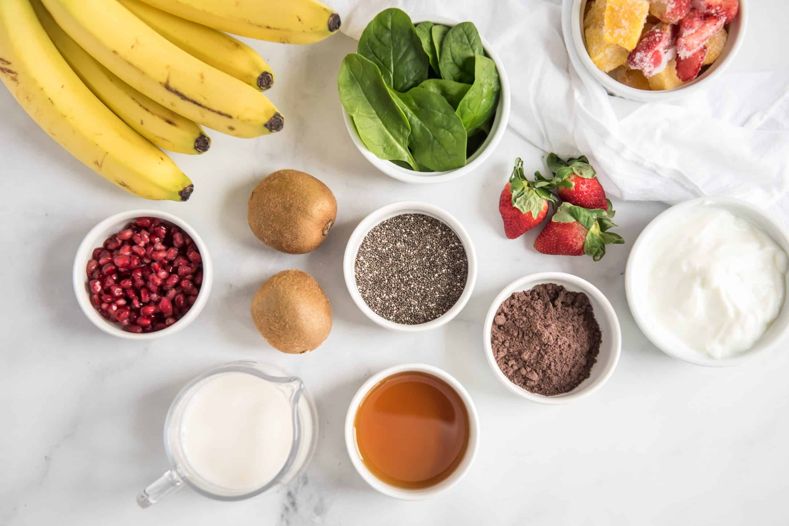 Bananas, baby spinach, kiwi fruit, almond milk, chia seeds, pomegranate seeds and frozen fruit on a white marble background.