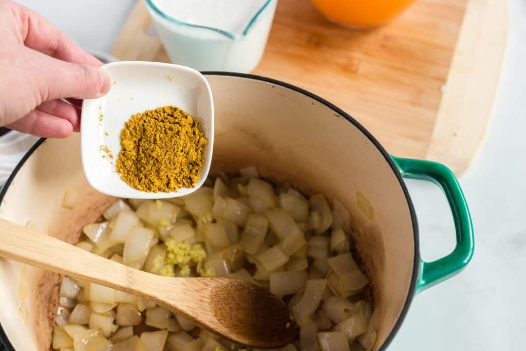 Curry being added to pot with cooked onions and wooden spoon.