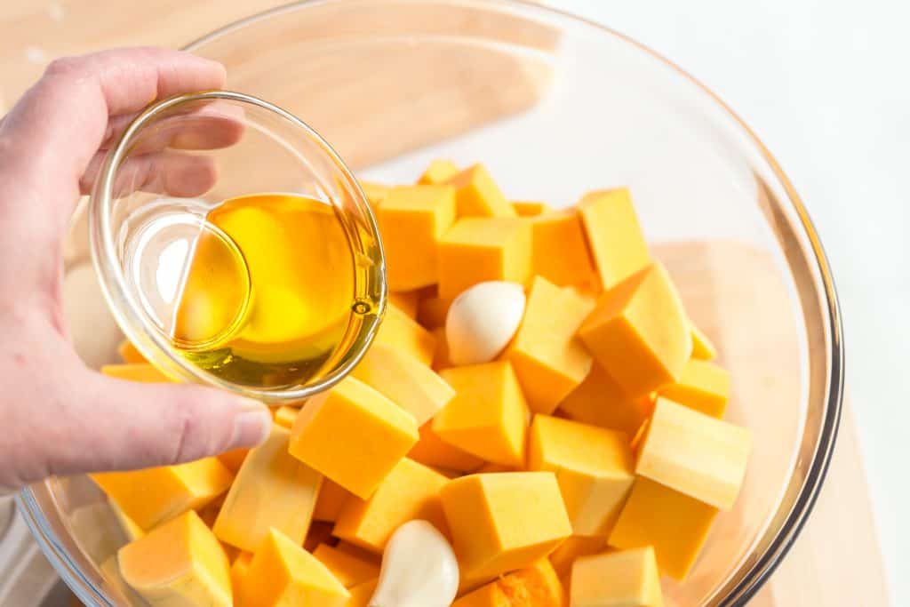 Butternut squash cut into chunks and garlic cloves in a glass bowl with olive oil being drizzled on top.