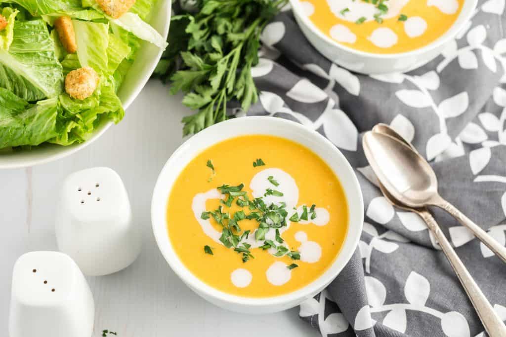 Vegan butternut squash soup topped with parsley and coconut milk on a white table with a black and white polo dot table cloth and two silver serving spoons. A Caesar salad and salt and pepper shakers are on the white table.