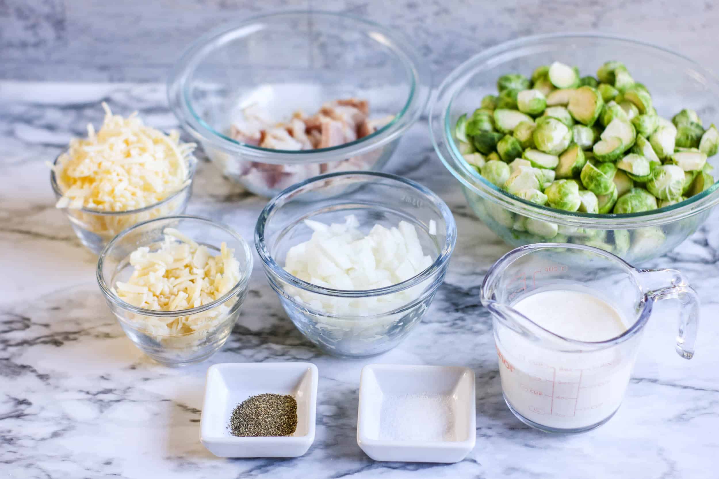 Brussels sprouts, cheese, onions, heavy cream, bacon, salt and pepper on a white marble background.