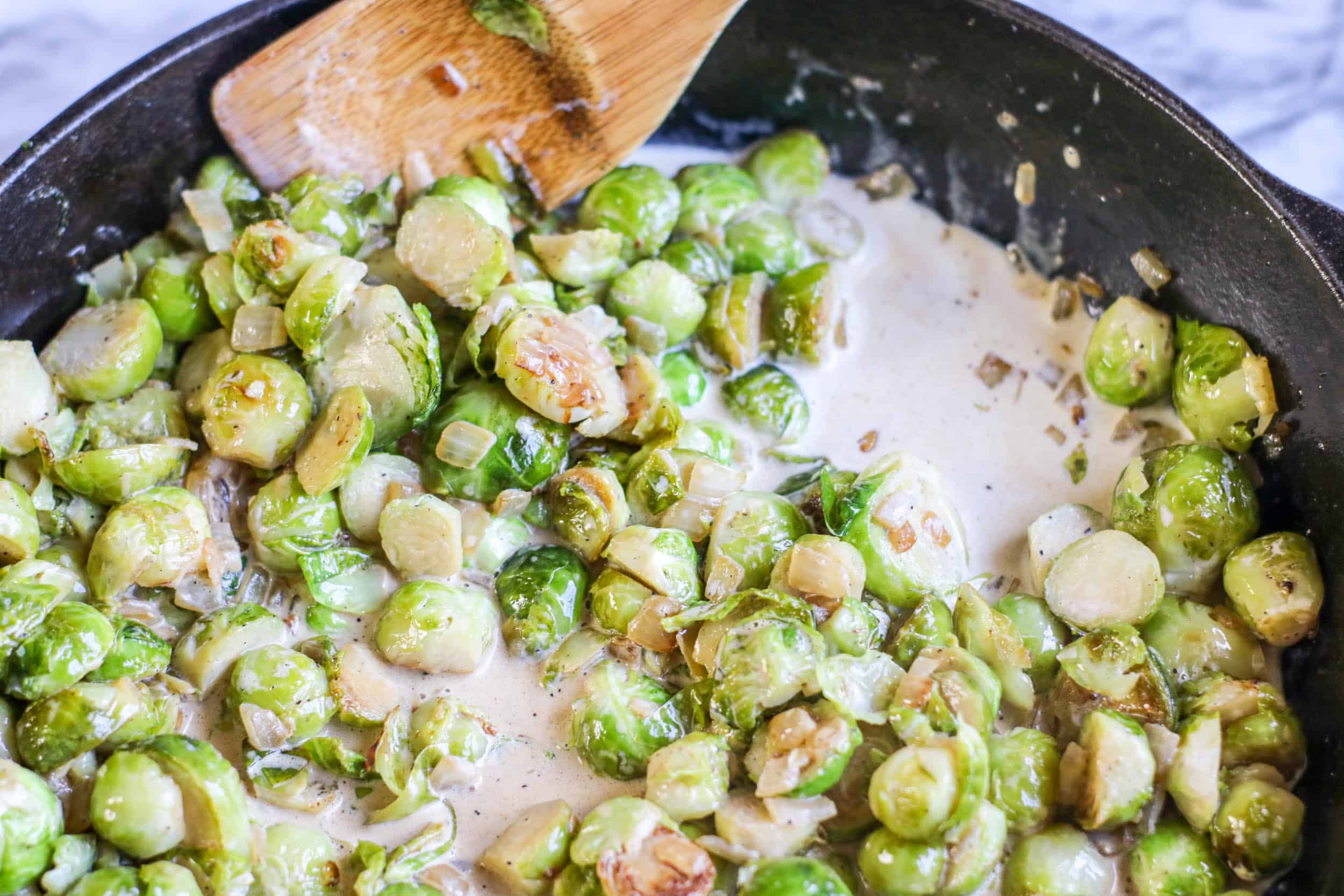 Brussels sprouts in a cast iron skillet with heavy cream and a wooden spoon.