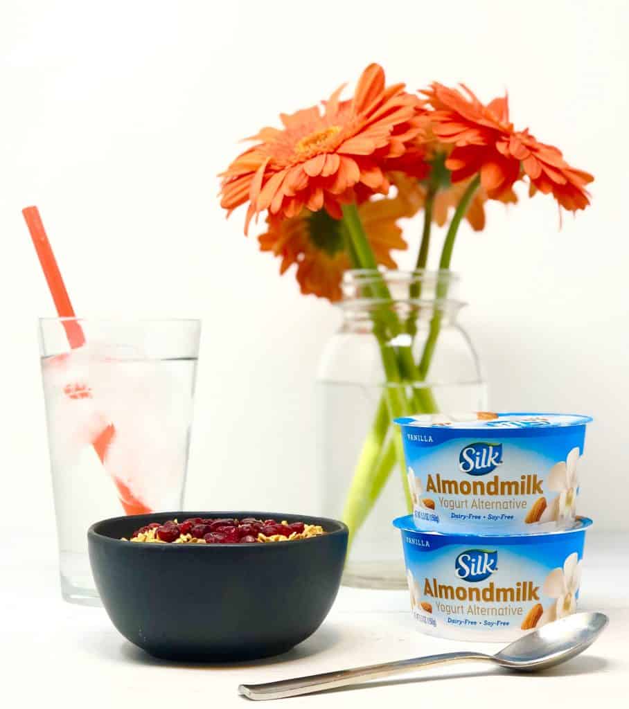 Smoothie bowl, glass of ice water, Silk almond yogurt and orange Gerber daisies on white background.