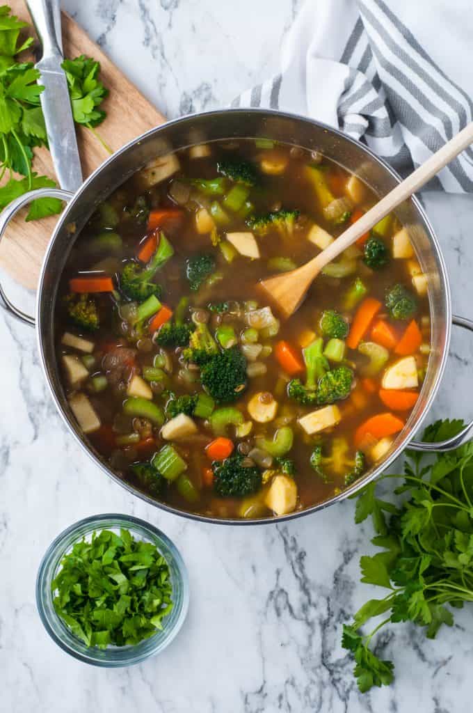 broccoli, carrots, celery, and parsnip in a pot with vegetable stock on a white background with parsley on the side.