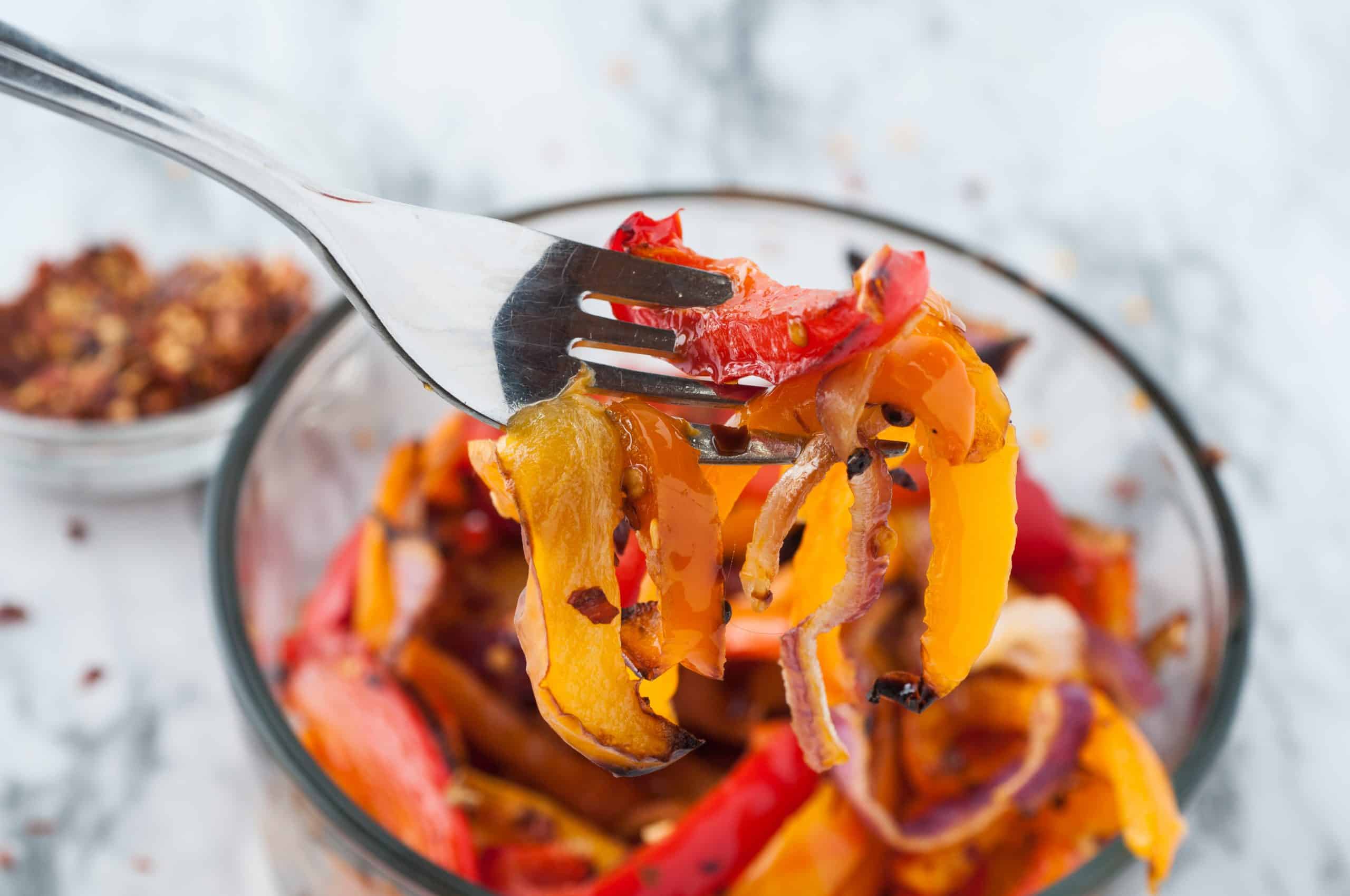 Close up of bell peppers and onions on a fork with crushed red pepper in a glass bowl in the background on white marble.