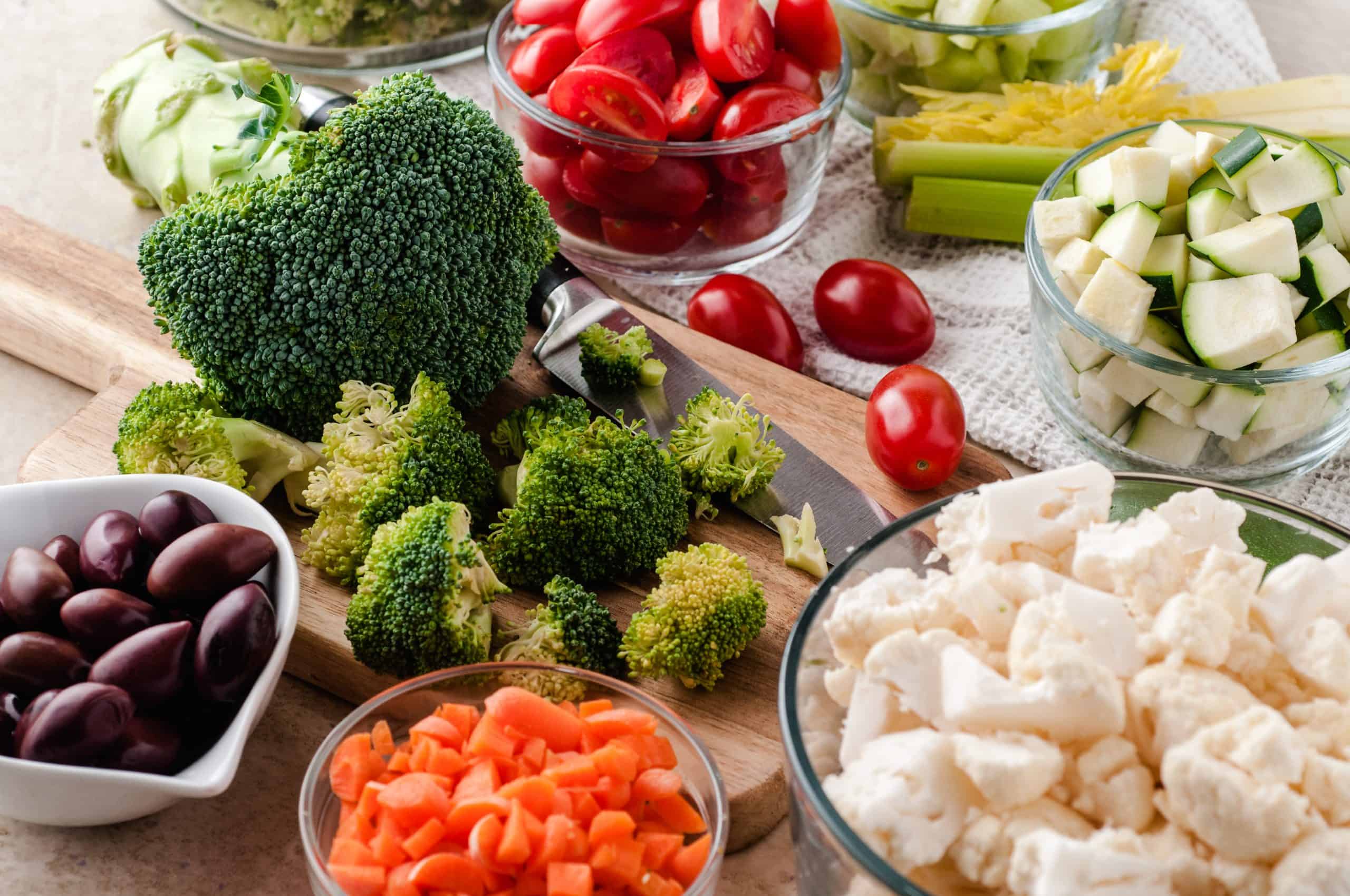 Fresh vegetables on a cutting board: broccoli, cauliflower, carrots, black olives, cherry tomatoes, zucchini and celery.