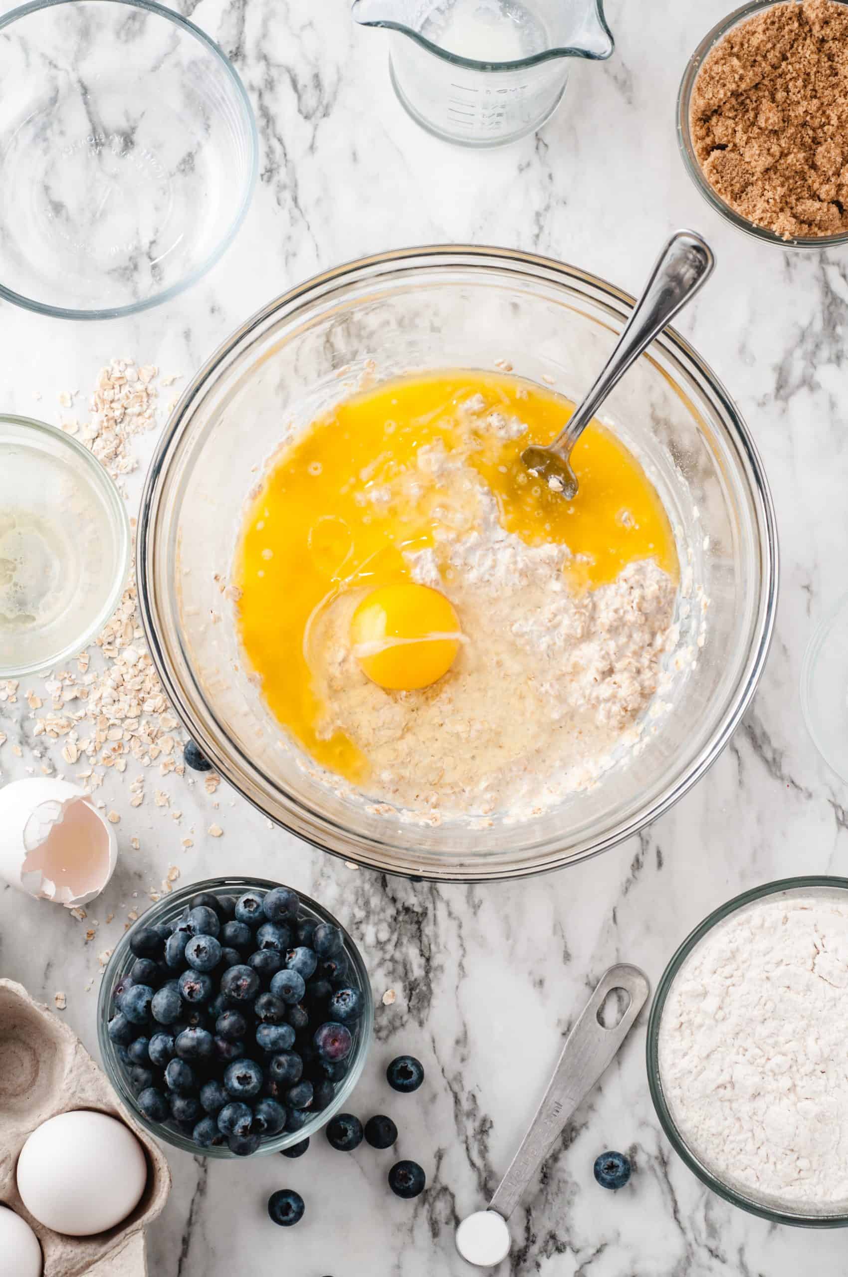 Oats, buttermilk egg and butter mixed in a bowl with blueberries, brown sugar and flour around the bowl on a white marble table.