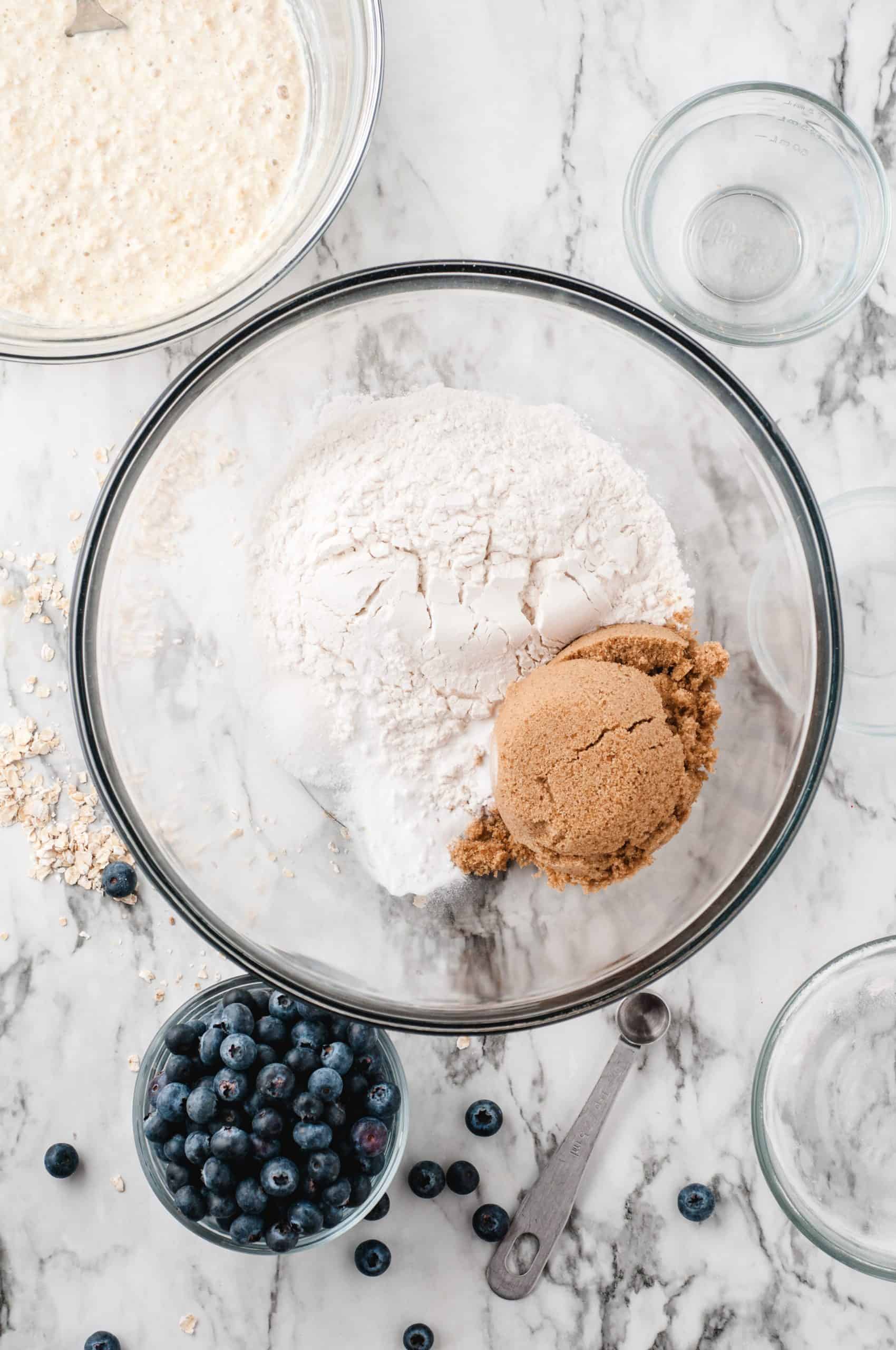Flour and brown sugar in a glass bowl with blueberries on the side.
