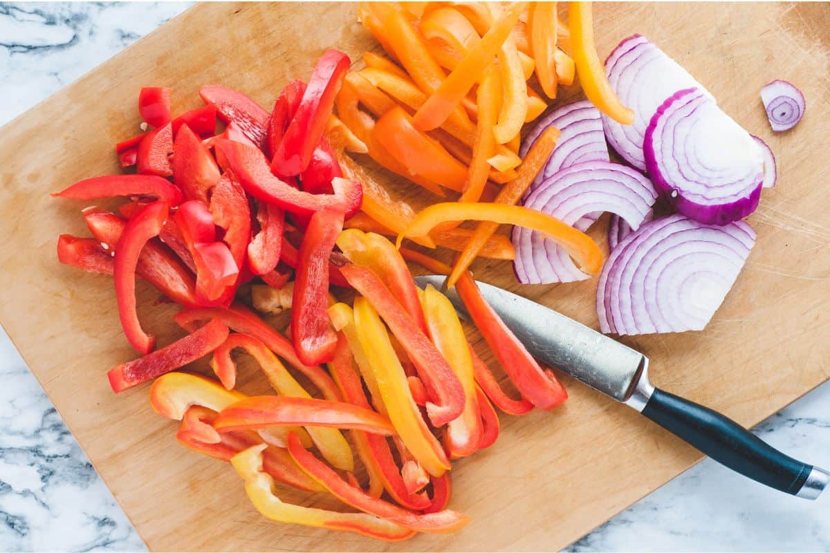 Overhead view of raw sliced bell peppers and onions on a wooden cutting board.