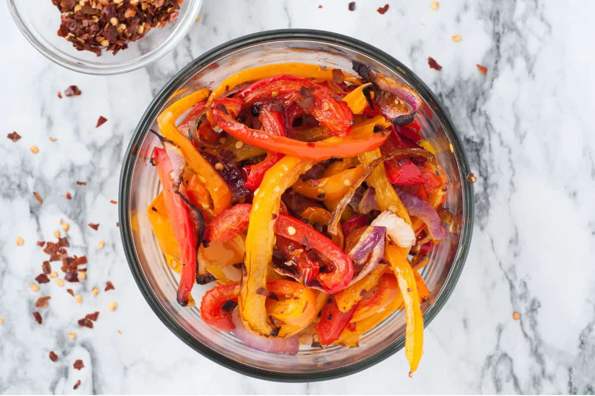 Overhead view of roasted bell peppers and onions with red pepper flakes in a glass bowl.