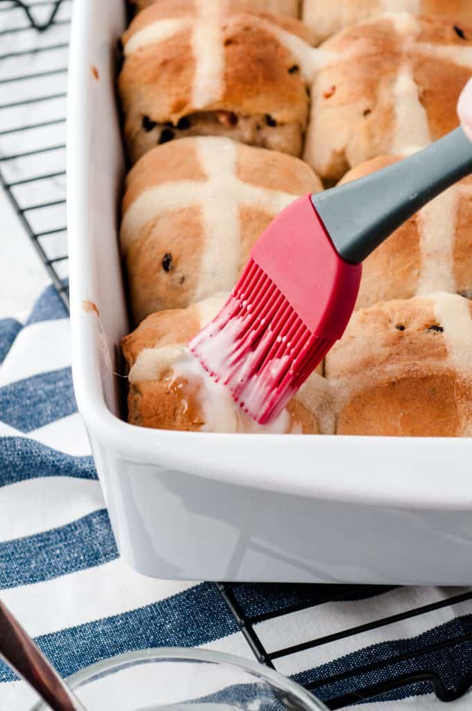 Icing being brushed onto buns in a baking pan on a wire rack.