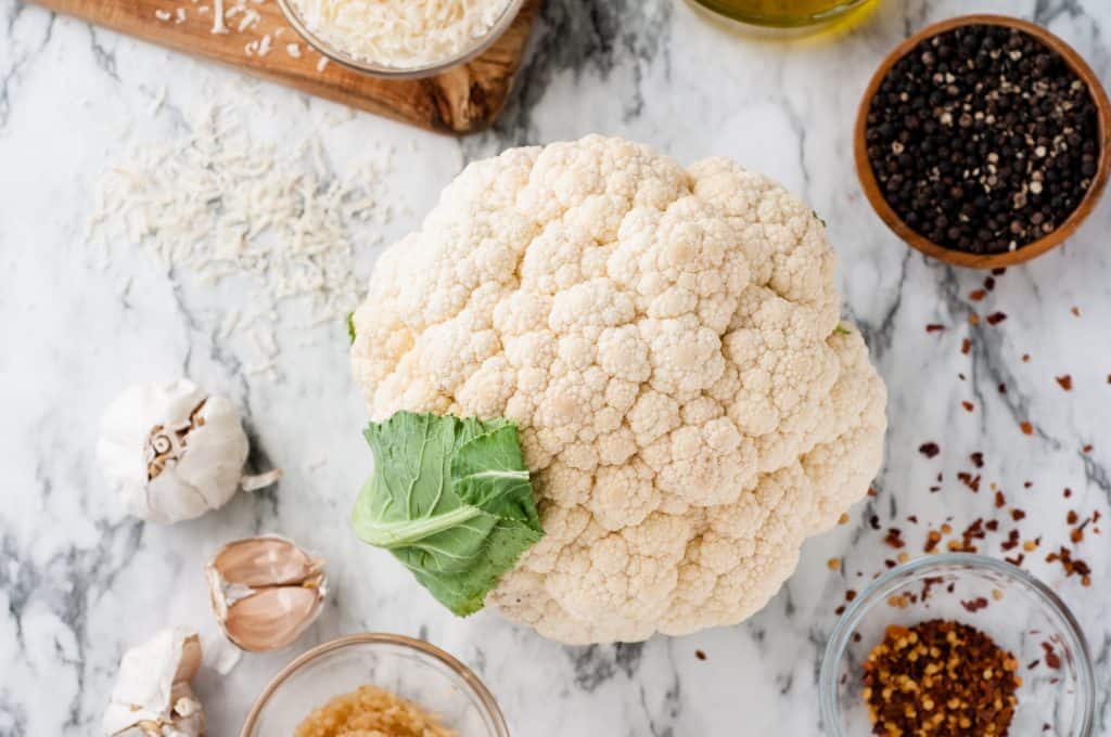Head of cauliflower on a white marble background with herbs, cheese and garlic around it.