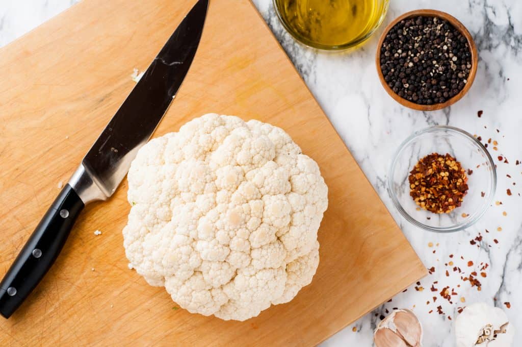 Head of cauliflower on a cutting board with large knife beside it and pepper, olive oil, and crushed red pepper on the side on a marble background.