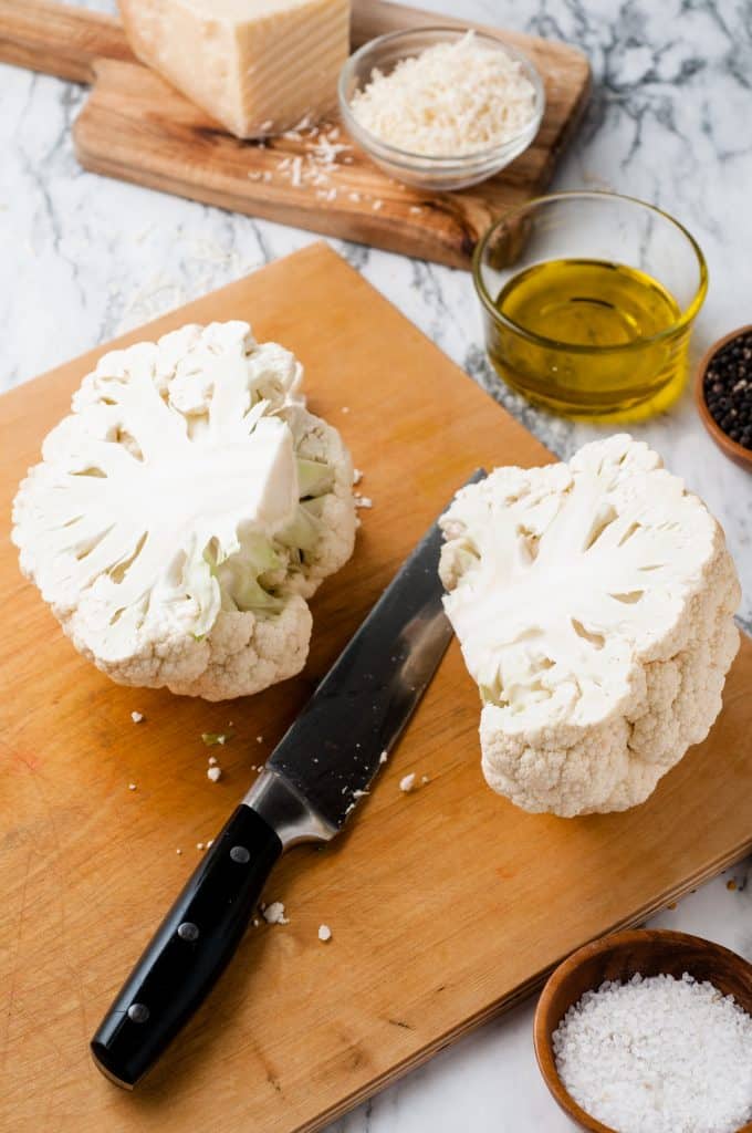 Cauliflower head sliced in half on a cutting board with a large knife.