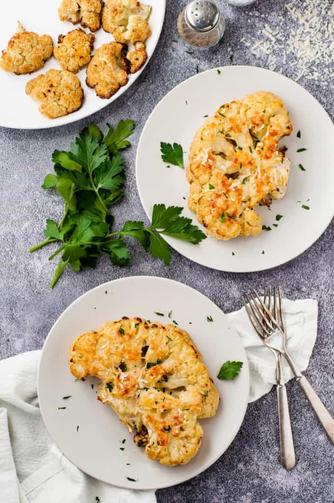 2 Roasted cauliflower steaks on white plates on a gray marble background with 2 forks on the side.