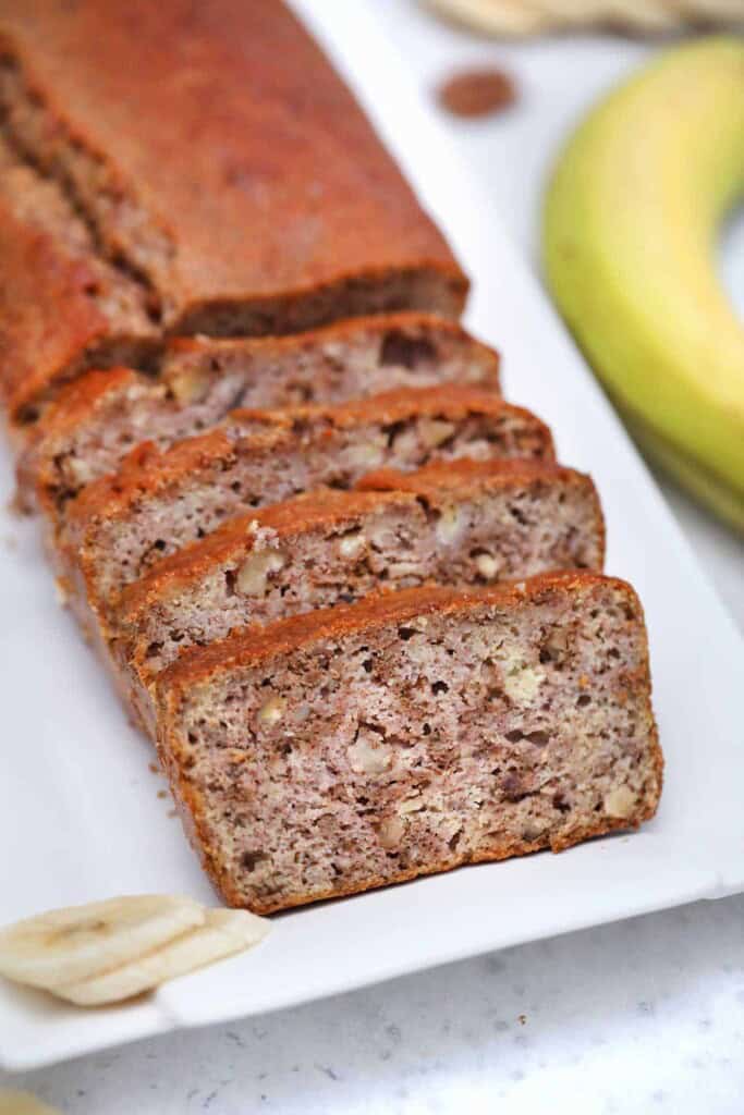 Side angle of sliced banana bread on a white platter with fresh banana garnish and banana on the side with a white background.
