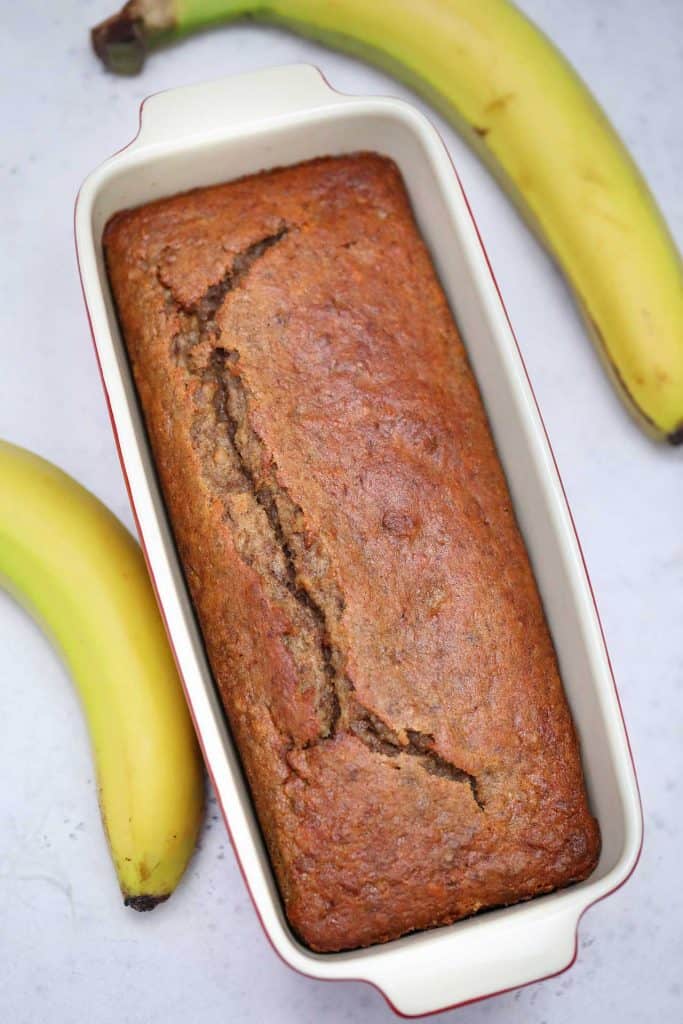 Baked banana bread in a loaf pan with a banana on each side on a white background. 