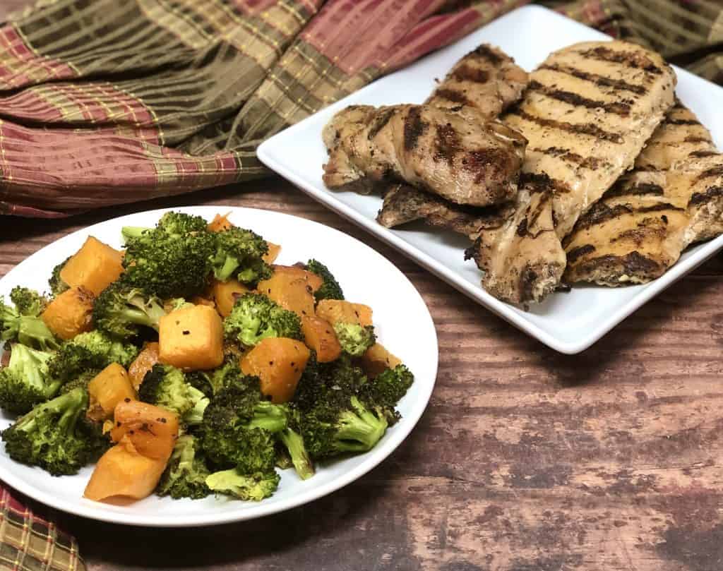 Roasted broccoli and sweet potatoes on a plate with grilled chicken behind it.