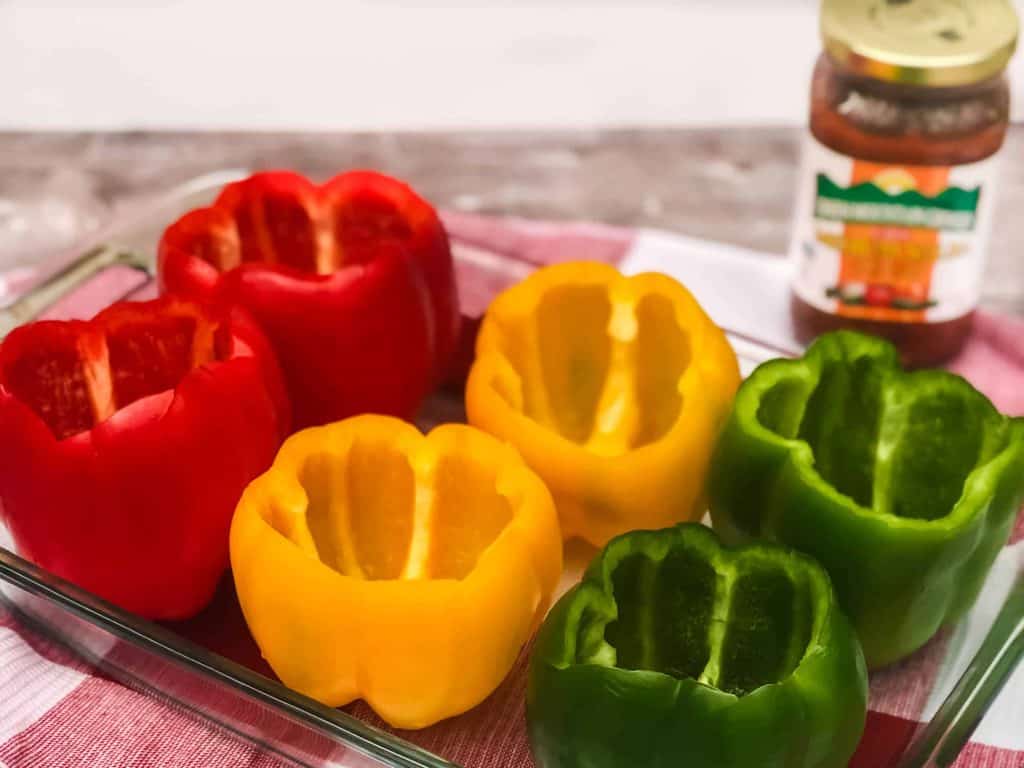 Red, yellow and green bell peppers hollowed out and seeded in baking dish with a jar of salsa behind them.