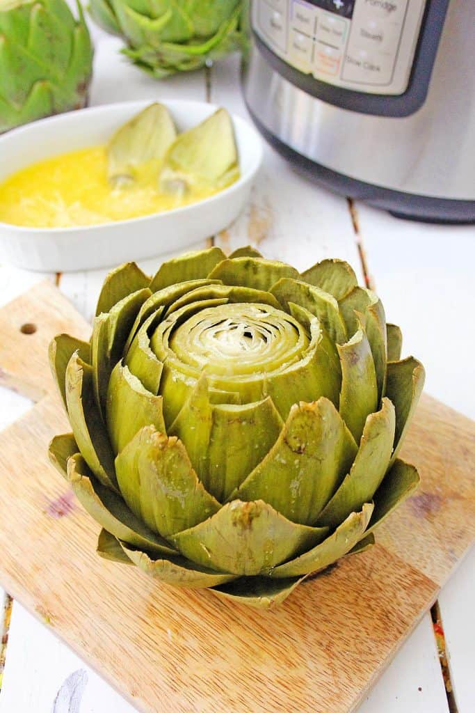 Instant Pot Artichokes on a cutting board with butter sauce and leaves of artichokes in it behind the cutting board and Instant Pot to the side.