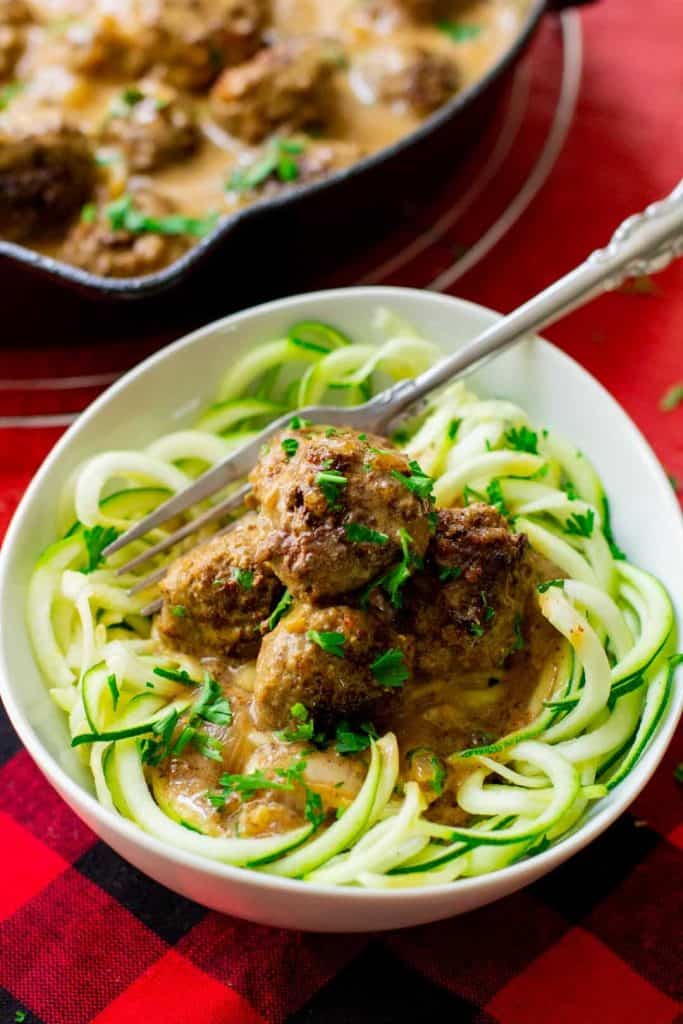 Keto Swedish meatballs over zucchini noodles in a white bowl with a fork in it and a black iron skillet with Swedish meatballs in it behind the bowl.