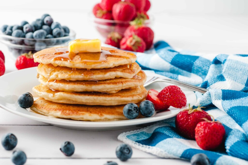 Stack of delicious pancakes with butter and maple syrup and fruit surrounding it with a blue and white checked kitchen towel.