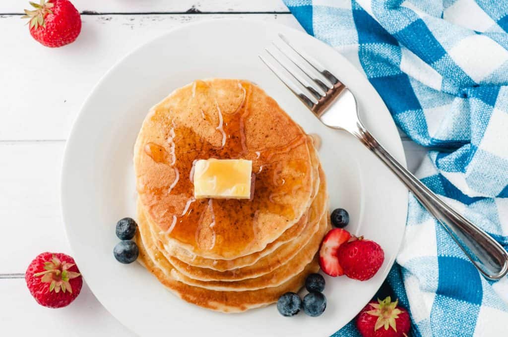 Overhead shot of pancakes on a white plate with a pat of butter and drizzle of maple syrup surrounded with strawberries and blueberries.
