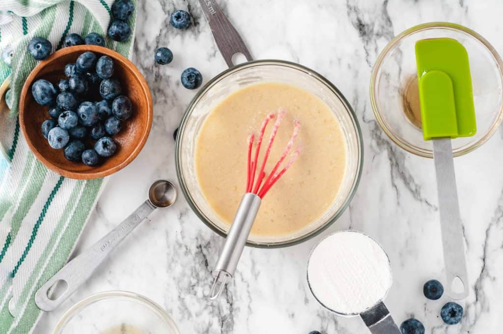 Blueberries, cake batter in glass bowl with whisk in it and cup of flour and green spatula on the side on a white marble background.
