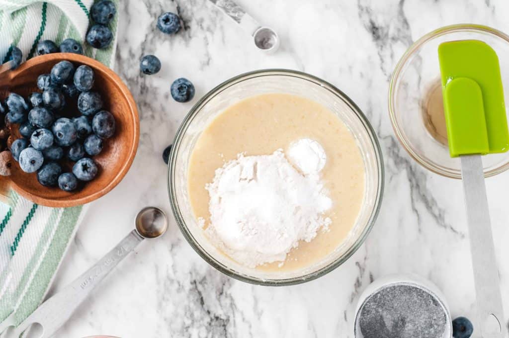 Flour on top of cake batter, not stirred in, blue berries on the side and green spatula on a marble background.