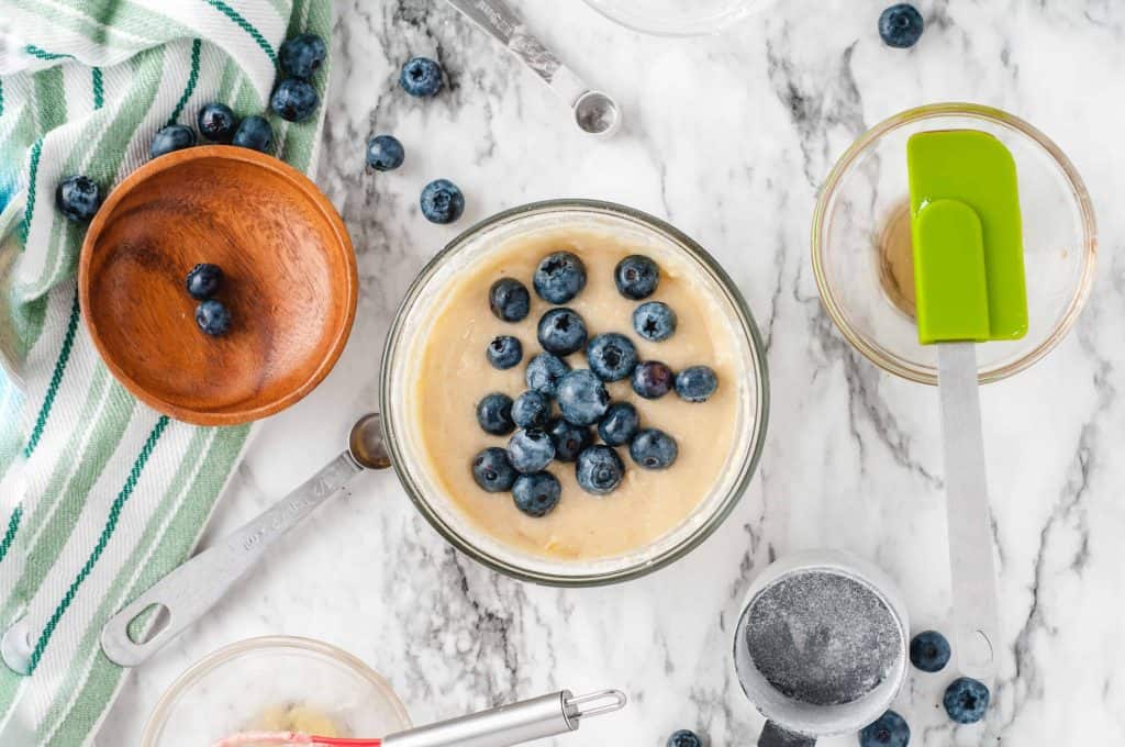 Blueberries on top of cake batter with wood bowl and green spatula on top of glass bowl.