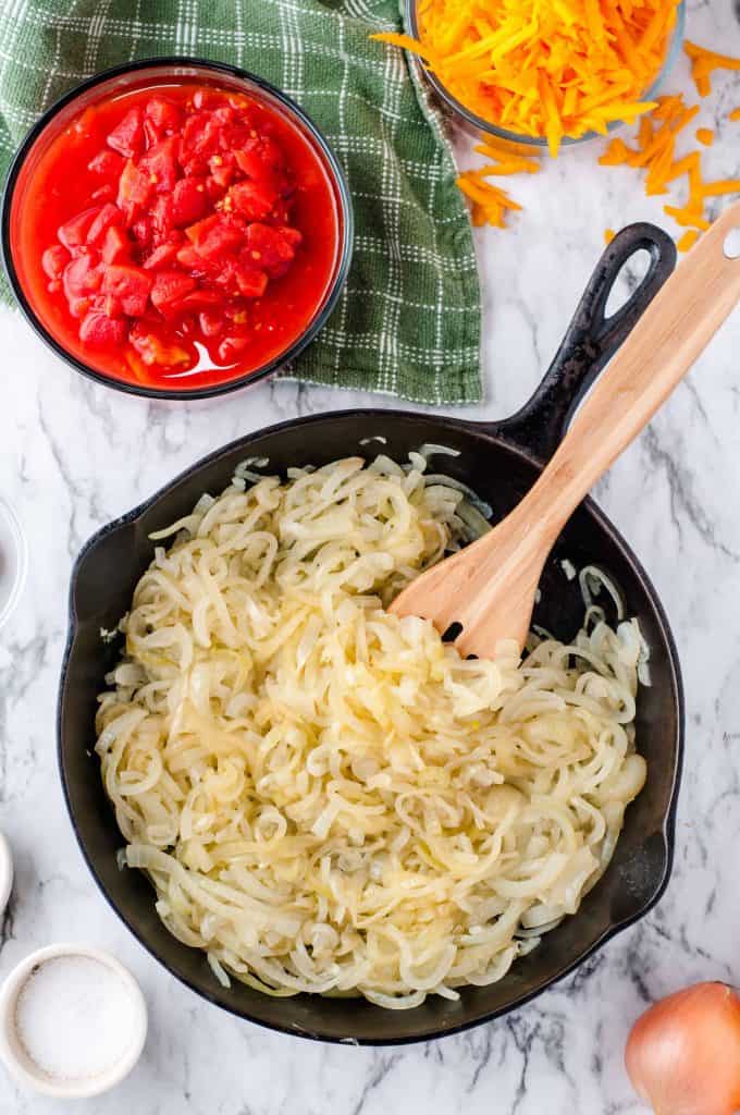 Cooked onions in a cast iron skilled with bowl of canned tomatoes in top left corner on green kitchen towel.