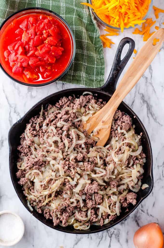 Beef and onions combined in cast iron skillet with a bowl of stewed tomatoes in top left hand corner on top of green kitchen towel.