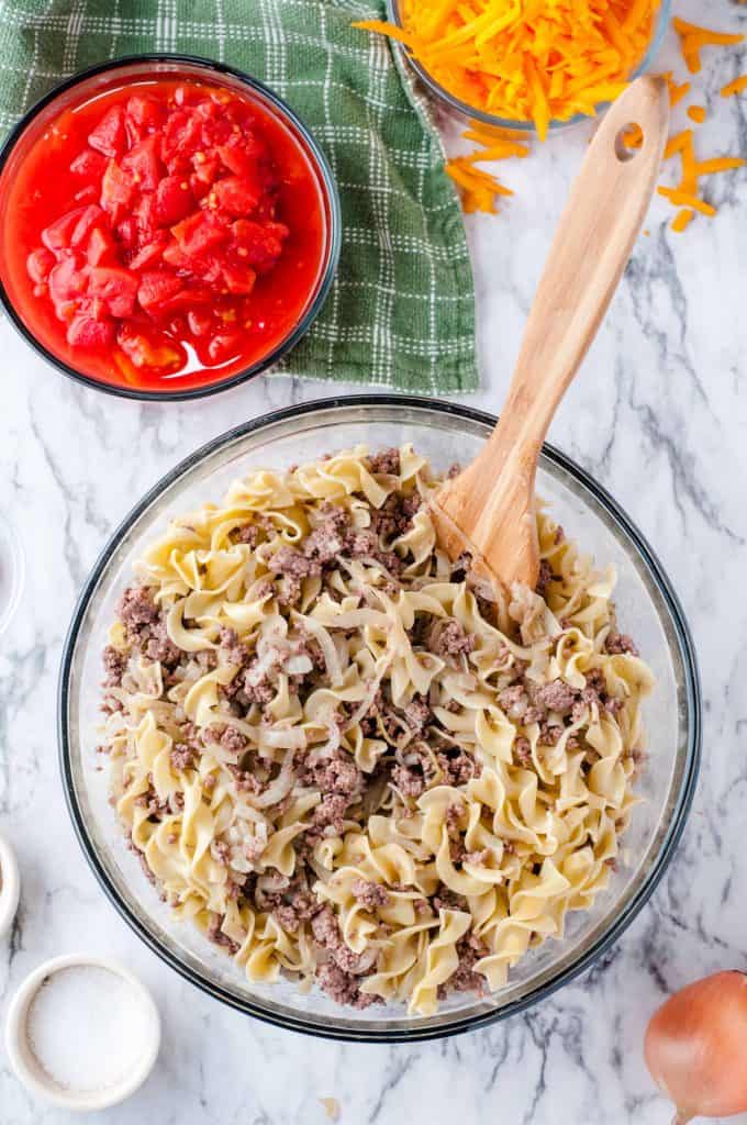 Beef and egg noodles combined in a large glass bowl for casserole with a bowl of canned diced tomatoes to side.