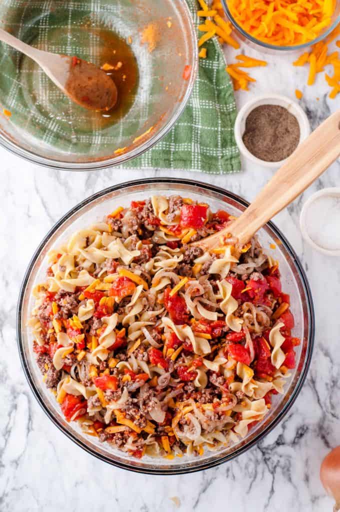 Cheesy beef and noodle casserole ingredients being stirred in a large glass bowl with a wooden spoon.