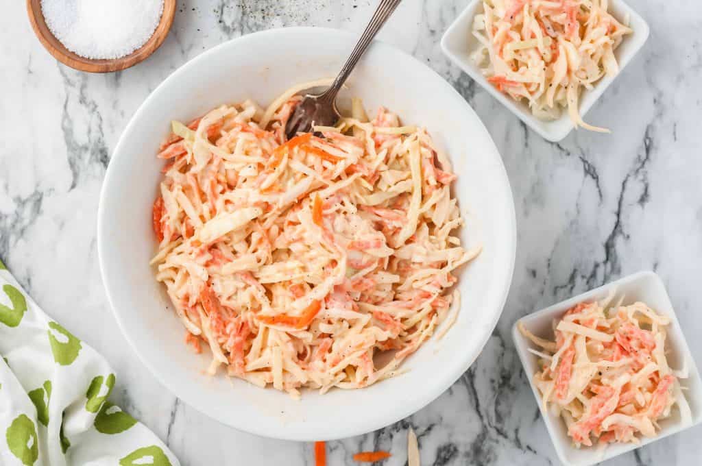 Classic Creamy Coleslaw in a white bowl with a salad fork in it and 2 servings in square white bowls on the side. A wooden bowl with coarse kosher sea salt in the upper left hand corner.