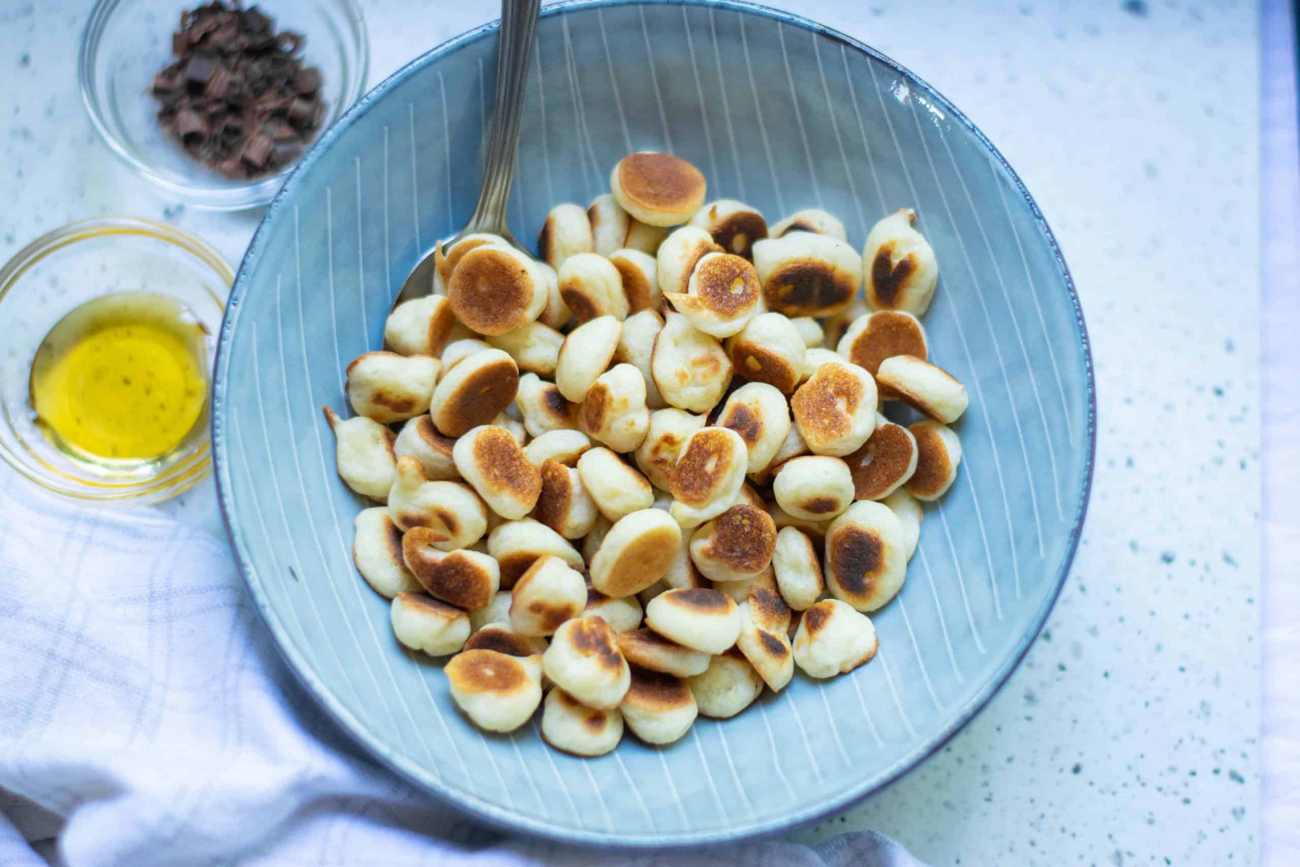 Overhead shot of mini pancakes cereal with a spoon in the bowl and cocoa nibs and agave nectar on the side.