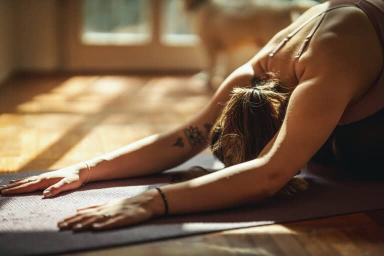 Young woman is doing yoga meditation in the living room at home. She is meditating on floor mat in morning sunshine.