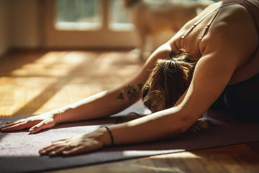 Young woman is doing yoga meditation in the living room at home. She is meditating on floor mat in morning sunshine.